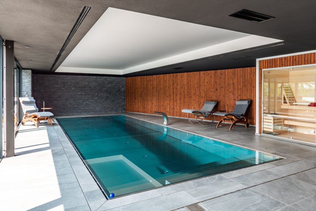 Moderner Indoorpool mit Liegen und angrenzender Sauna in elegantem Wellnessbereich mit Holz- und Steinwänden.