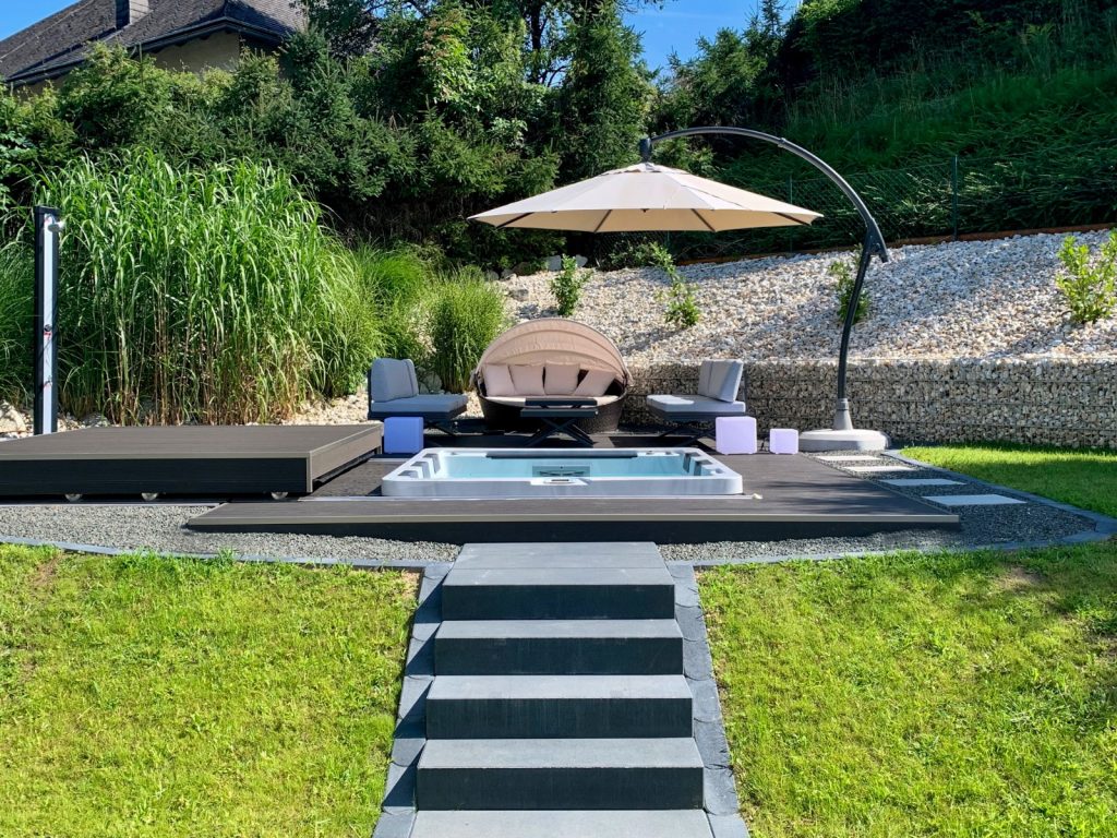 Modern gestalteter Garten mit einer Treppe zu einem eingelassenem Whirlpool, Sonnenliegen und Sonnenschirm.