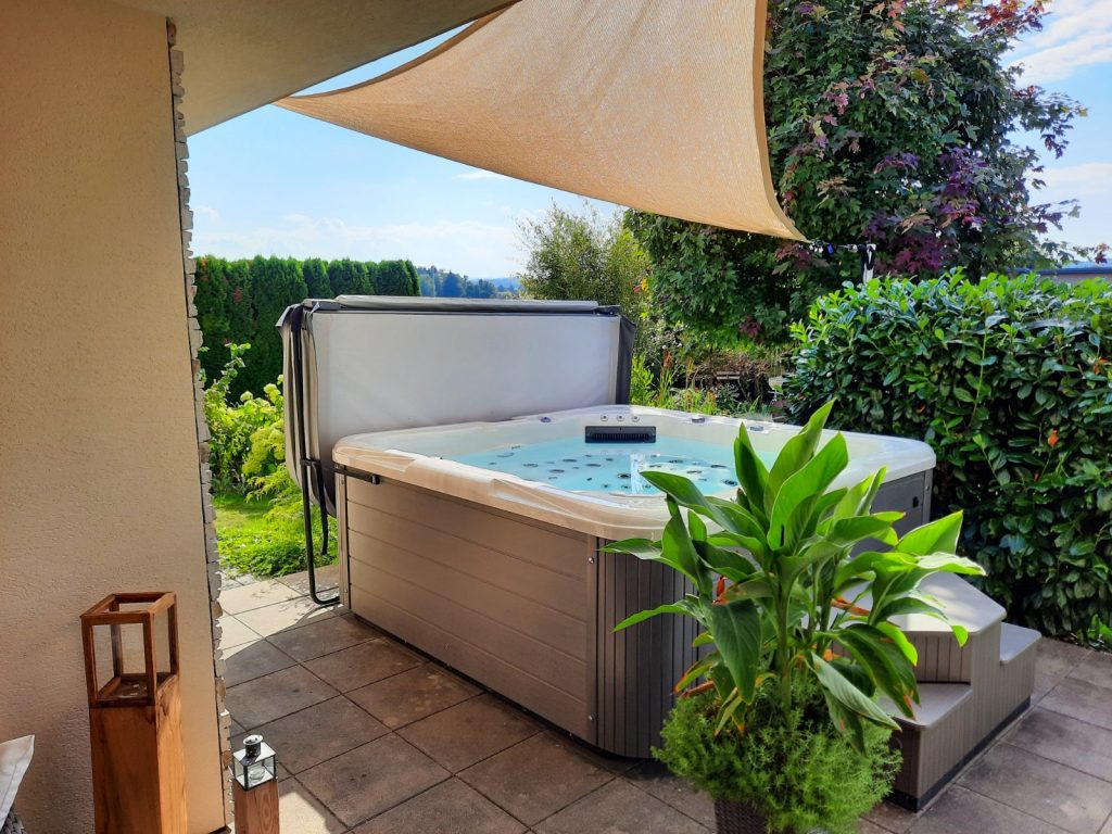 Whirlpool auf Terrasse mit Sonnensegel und Pflanzen, eingebettet in grünen Garten mit Hecken und Bäumen.