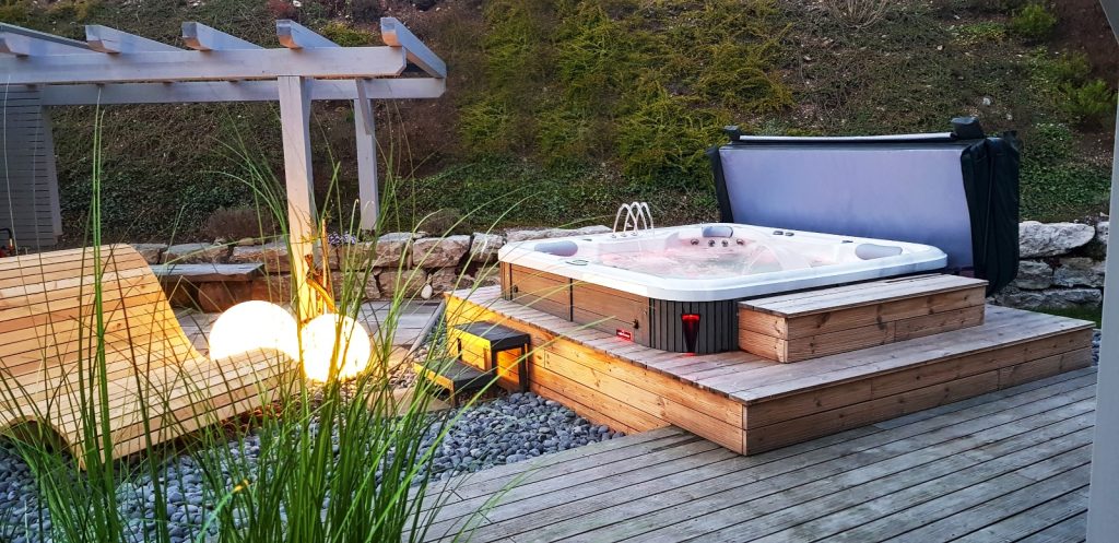 Whirlpool auf Holzpodest mit stimmungsvoller Beleuchtung und moderner Liege im Garten bei Abenddämmerung.