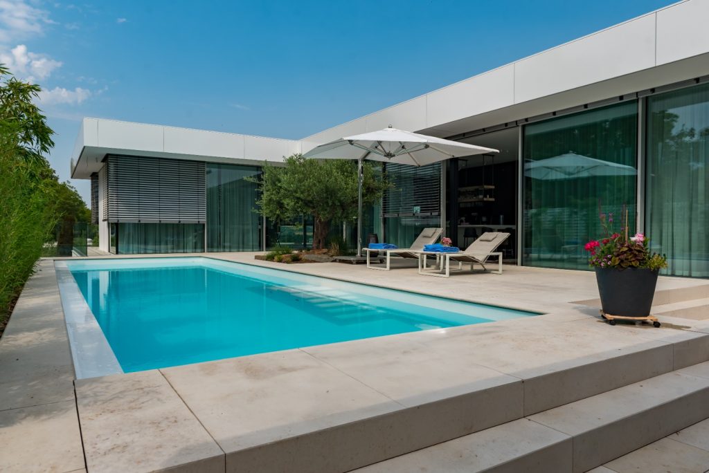 Moderner Pool mit Liegen und Sonnenschirm vor einem minimalistischen Haus mit Glasfront und Olivenbaum.