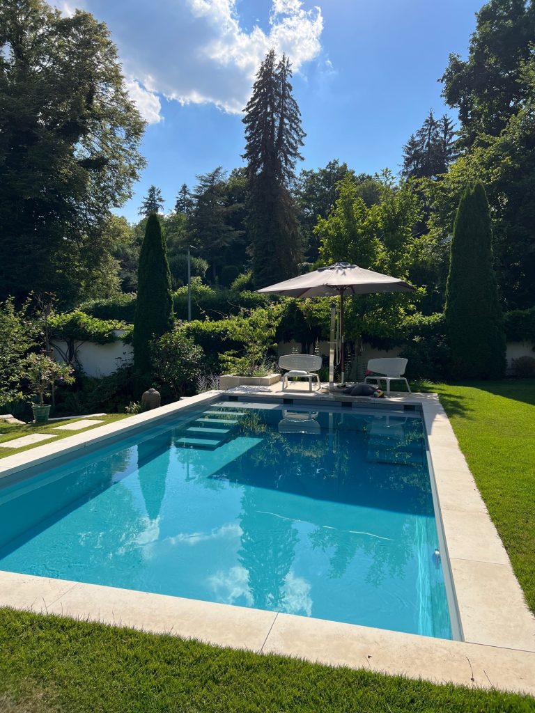 Gartenpool mit Sonnenschirm und Liegen, umgeben von Bäumen und Rasen bei strahlendem Sommerwetter.