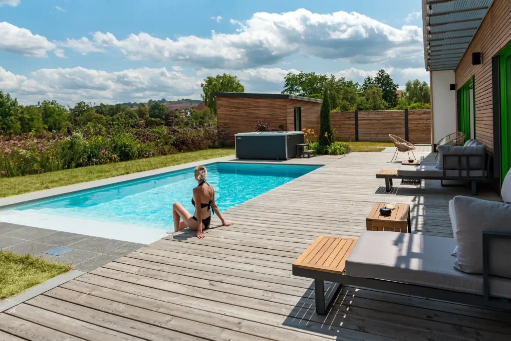 Frau sitzt am Rand eines modernen Gartenpools mit Holzterrasse und genießt die Sonne an einem Sommertag.