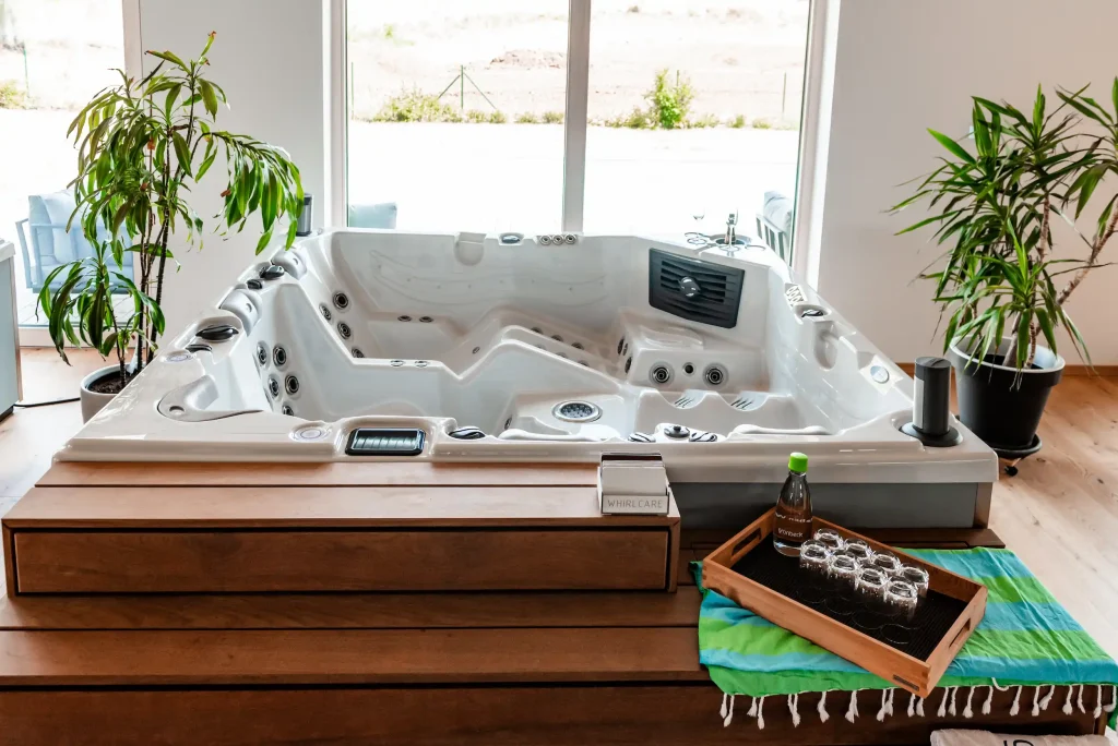 Moderner Whirlpool mit Liege- und Sitzplätzen im Innenraum mit Holzstufen, Pflanzen und Tablett mit Gläsern und Mineralwasser.