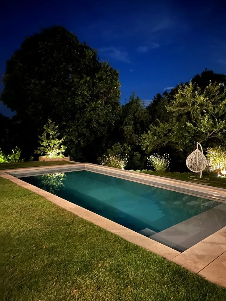 Beleuchteter Gartenpool bei Nacht, umgeben von Bäumen und Rasen, mit modernem Hängesessel im Hintergrund.