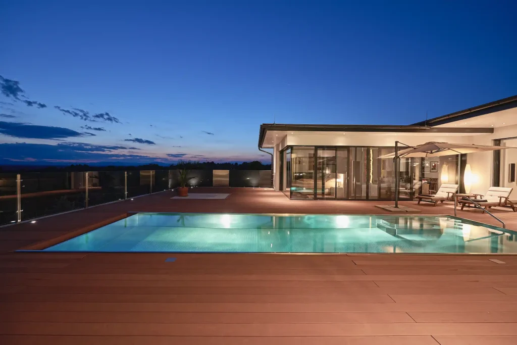 Beleuchteter Pool auf einer Terrasse bei Abenddämmerung vor einem modernen Haus mit Glasfassade.