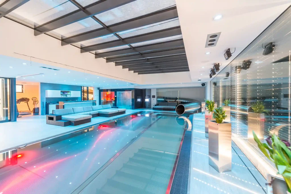 Luxuriöser Indoor-Pool mit moderner Beleuchtung, Lounge-Bereich und Tunnelrutsche aus Edelstahl in elegantem Design.