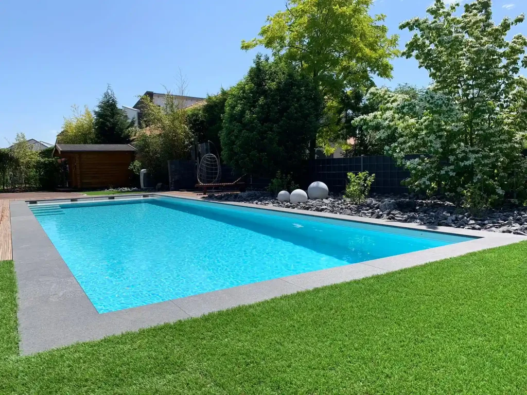 Eleganter Gartenpool mit grauer Randverkleidung, gepflegtem Rasen und moderner Gartengestaltung mit Natursteinen und Bäumen.