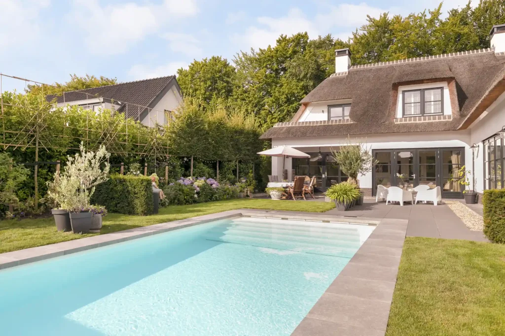 Hochwertiger Gartenpool mit grauer Umrandung vor einem stilvollen Haus mit Reetdach und Terrasse mit Sitzmöglichkeiten.