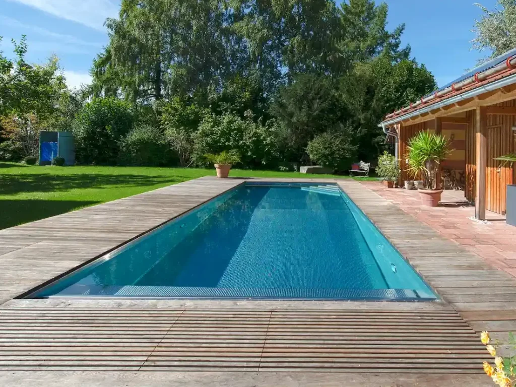 Langer, rechteckiger Gartenpool mit Holzumrandung neben einer Terrasse und begrüntem Garten mit Bäumen.