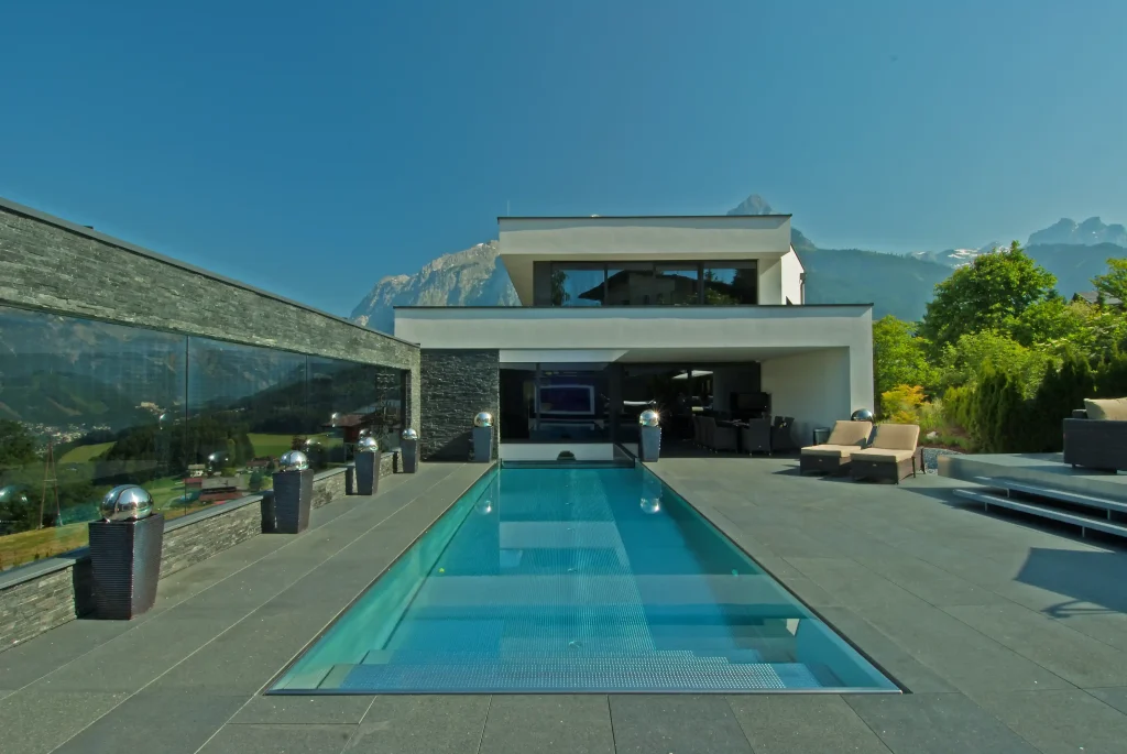 Modernes Haus mit Infinity-Pool und Bergblick, umgeben von einer Terrasse mit grauen Steinfliesen und Sonnenliegen.
