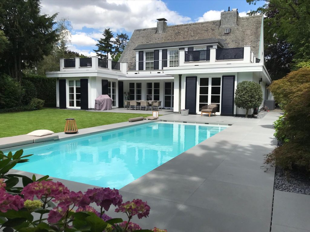 Moderner Garten mit rechteckigem Pool vor einem weißen Einfamilienhaus mit Terrasse und Rasenfläche.
