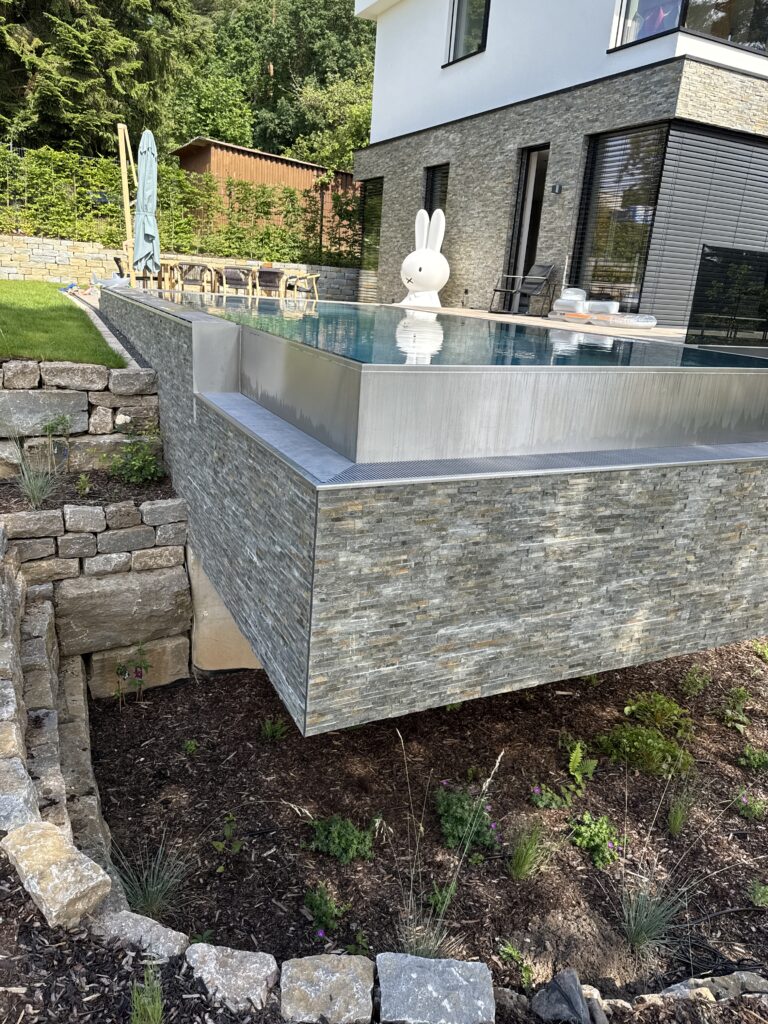 Tieferliegende Überlaufrinne und Infinity-Kante des Gartenpools aus Edelstahl. Im Hintergrund ist eine Figur und das Wohnhaus mit angrenzender Sitzecke zu sehen. Der Pool ist umgeben von Rasen und einem Beet aus Blumen und Steinen.