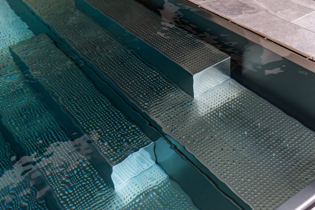 Detailaufnahme eines modernen Edelstahlpools mit integrierter Treppenanlage und gelochten Antirutschstufen. Das klare Wasser umspielt die präzise gefertigten Stufen und unterstreicht das hochwertige, funktionale Design im minimalistischen Stil.