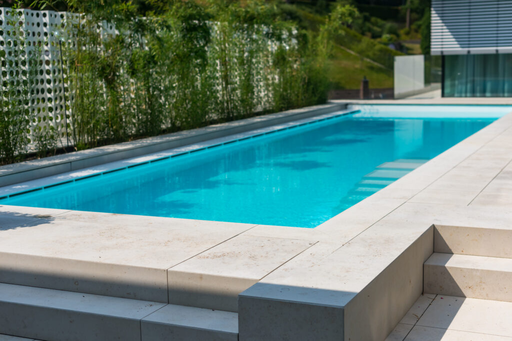Moderner, länglicher Außenpool mit ruhigem Wasserspiegel, eingebettet in eine großzügige Naturstein-Terrasse. Stilvolle Liegen, Sonnenschirm und ein dekorativer Sichtschutz aus Pflanzen schaffen eine entspannte, hochwertige Wohlfühlatmosphäre im Garten.