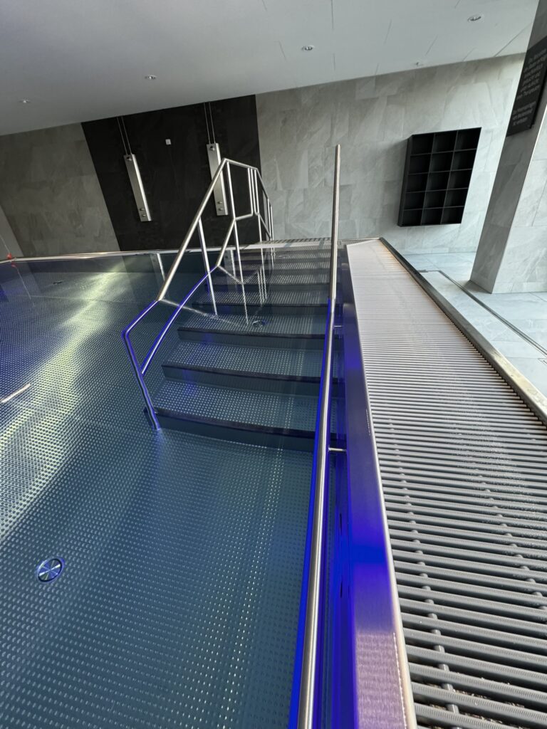 Moderner Indoor‑Pool als Therapiebecken mit großzügiger Treppenanlage für barrierearmen Einstieg. Die rutschhemmenden Stufen und die beidseitigen Edelstahlhandläufe bieten höchste Sicherheit und Komfort für Patienten. Die umlaufende Überlaufrinne sowie die dezente Beleuchtung unterstreichen das hochwertige Design und schaffen optimale Bedingungen für therapeutische Anwendungen.