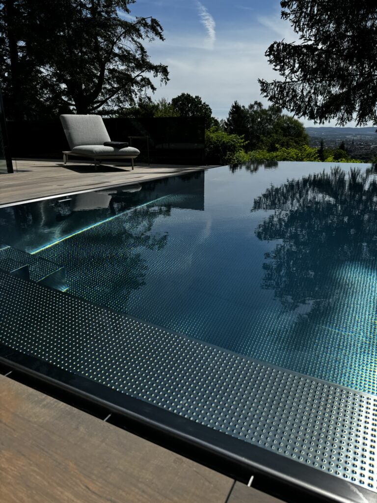 Moderner Infinity-Pool mit ruhigem Wasserspiegel und integrierter Überlaufrinne, harmonisch in eine stilvolle Terrasse mit hochwertiger Liege und Blick in die umliegende Landschaft eingebettet.