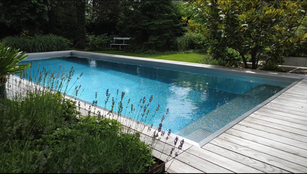 Modernes, rechteckiges Skimmerbecken aus Edelstahl, eingebettet in einen liebevoll gestalteten Garten. Der Pool ist von einer hellen, dezenten Beckenumrandung eingefasst und wird auf einer Seite von einer großzügigen Holzterrasse begleitet, die als Zugang und Aufenthaltsbereich dient.