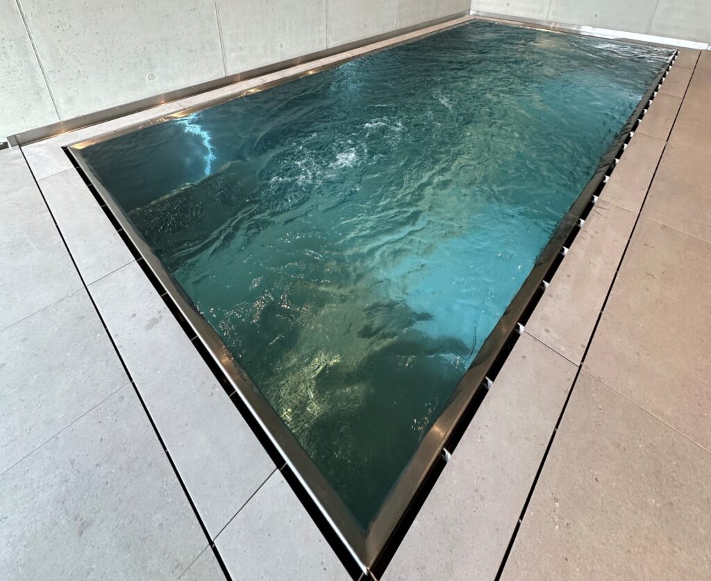 Modernes Indoor‑Becken mit klarer, rechteckiger Form und umlaufender Überlaufrinne. Die schlichte Architektur mit dezent integrierter Beleuchtung wird durch das bewegte Wasserbild betont und schafft eine ruhige, minimalistische Atmosphäre im Innenraum.