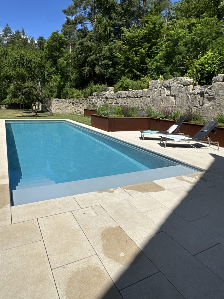Gartenpool aus GFK mit seitlicher Blocktreppe und verstecktem Rollabdeckungskasten. Sonnenliegen vor modernem Hochbeet runden die sommerliche Stimmung mit Sonnenlicht ab.
