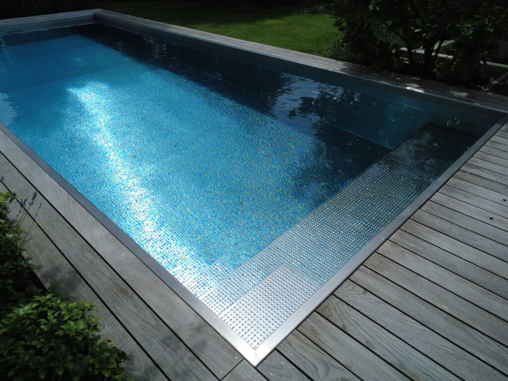 Blick auf den Gartenpool aus Edelstahl mit seitlicher Blocktreppe und umrahmender Holzterrasse.
Das Sonnenlicht spiegelt sich im Poolwasser.