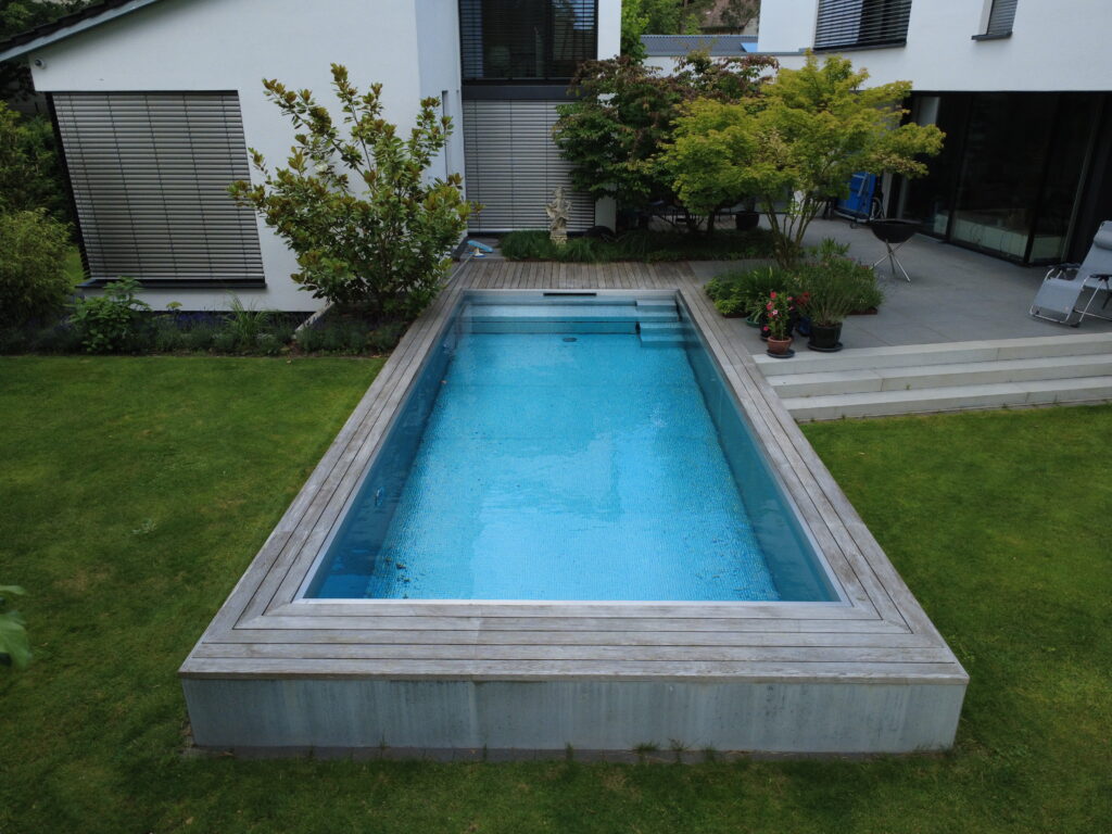 Edelstahlbecken mit Holzumrandung und Skimmer. Im Hintergrund Haus.