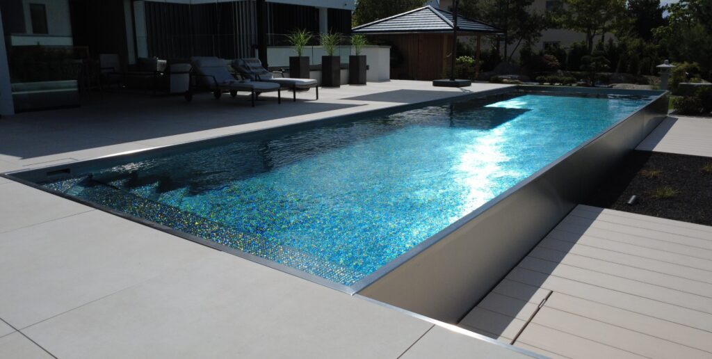 Blick von der Terrasse über die glitzernde Wasseroberfläche. Im Hintergrund des Pools befinden sich Sonnenliegen, die im Schatten stehen. Am Ende des Grundstücks steht ein Pavillon aus Holz.