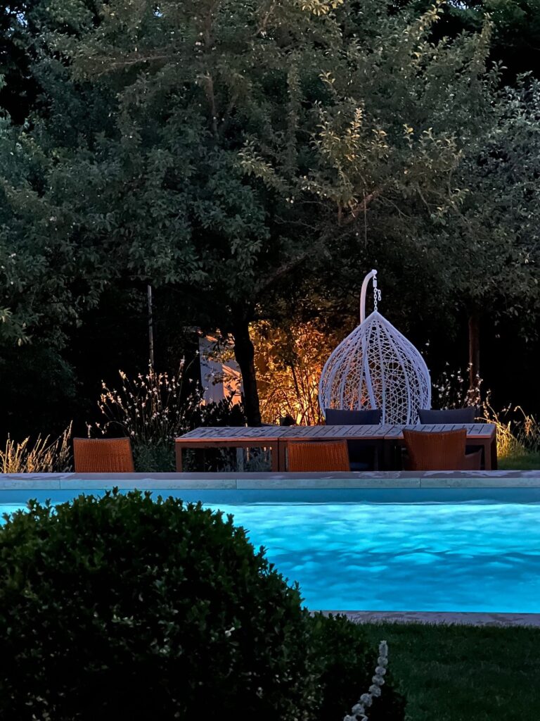 Gartenpool bei Abendlicht mit Esstisch und Stühlen im Hitergrund  umgeben von Bäumen und Rasen.