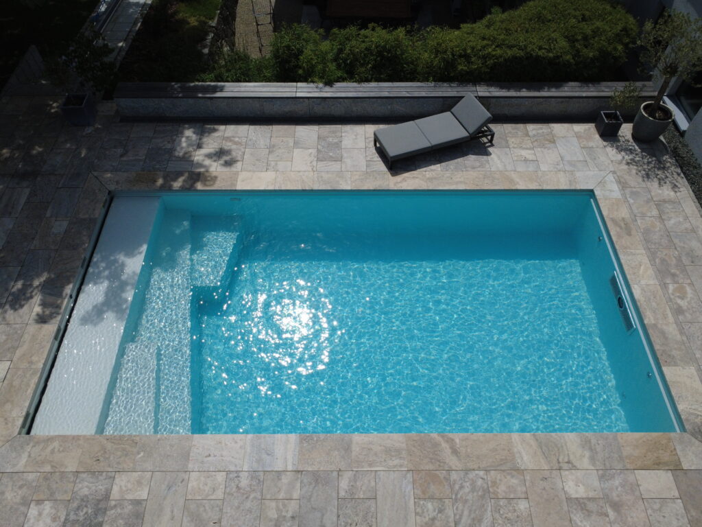 Blick auf das GFK-Becken mit Oblique-Treppe an der linken und Gegenstromanlage an der rechten Seite des Beckens.
Hinter dem Pool steht eine Liege